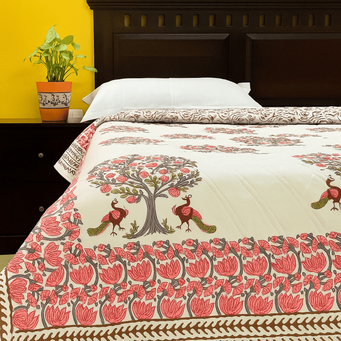 Cotton quilt with pink peacock and pomegranate tree block print, styled on a bed with white pillows and yellow background