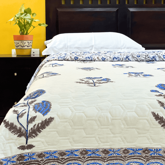 Handblock printed quilt with blue floral motifs and brown leafy detailing, laid on a bed with a dark wooden headboard and white pillows.