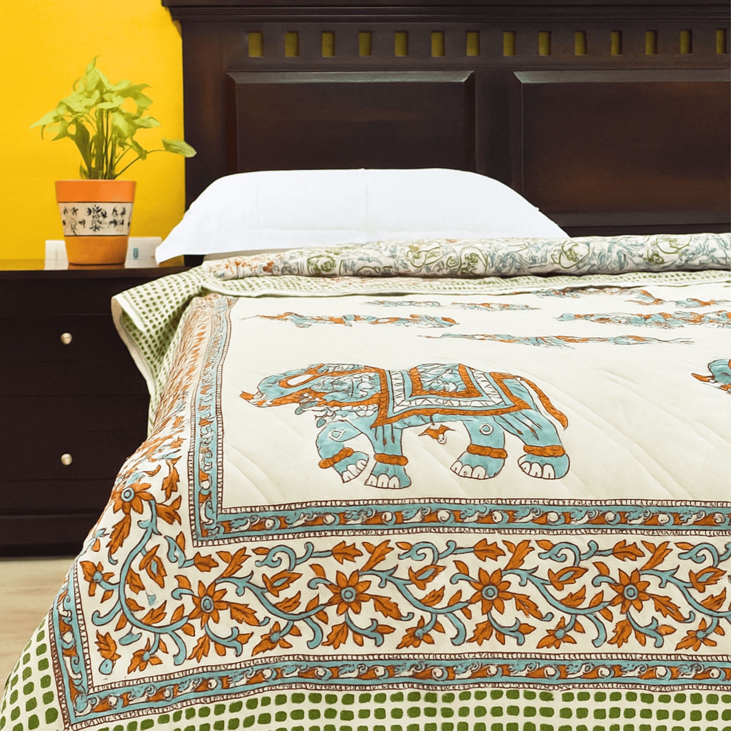 Handprinted cotton quilt with turquoise elephant motifs and floral borders, neatly spread on a bed with a wooden headboard.