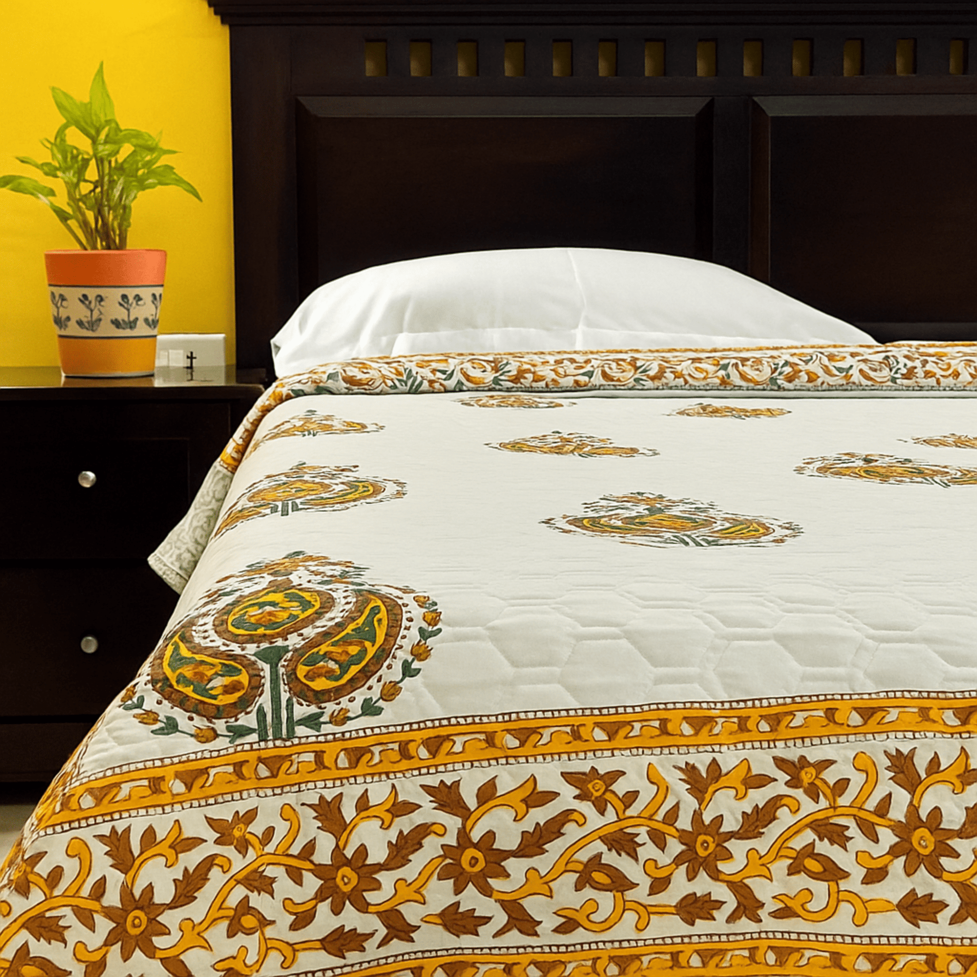 Handblock printed cotton quilt with traditional paisley motifs and floral yellow border, styled on a bed with white pillows and a dark wooden headboard.
