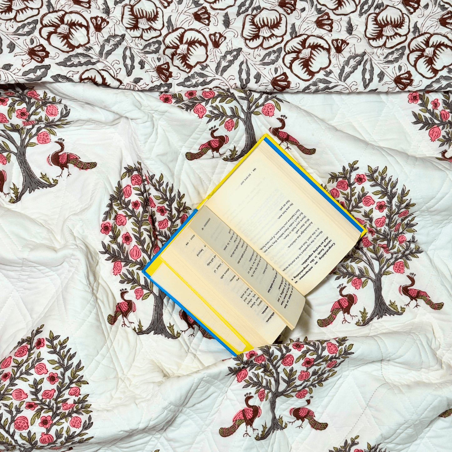Flat lay of block printed quilt with pink peacock tree motifs and brown floral print, styled with an open book