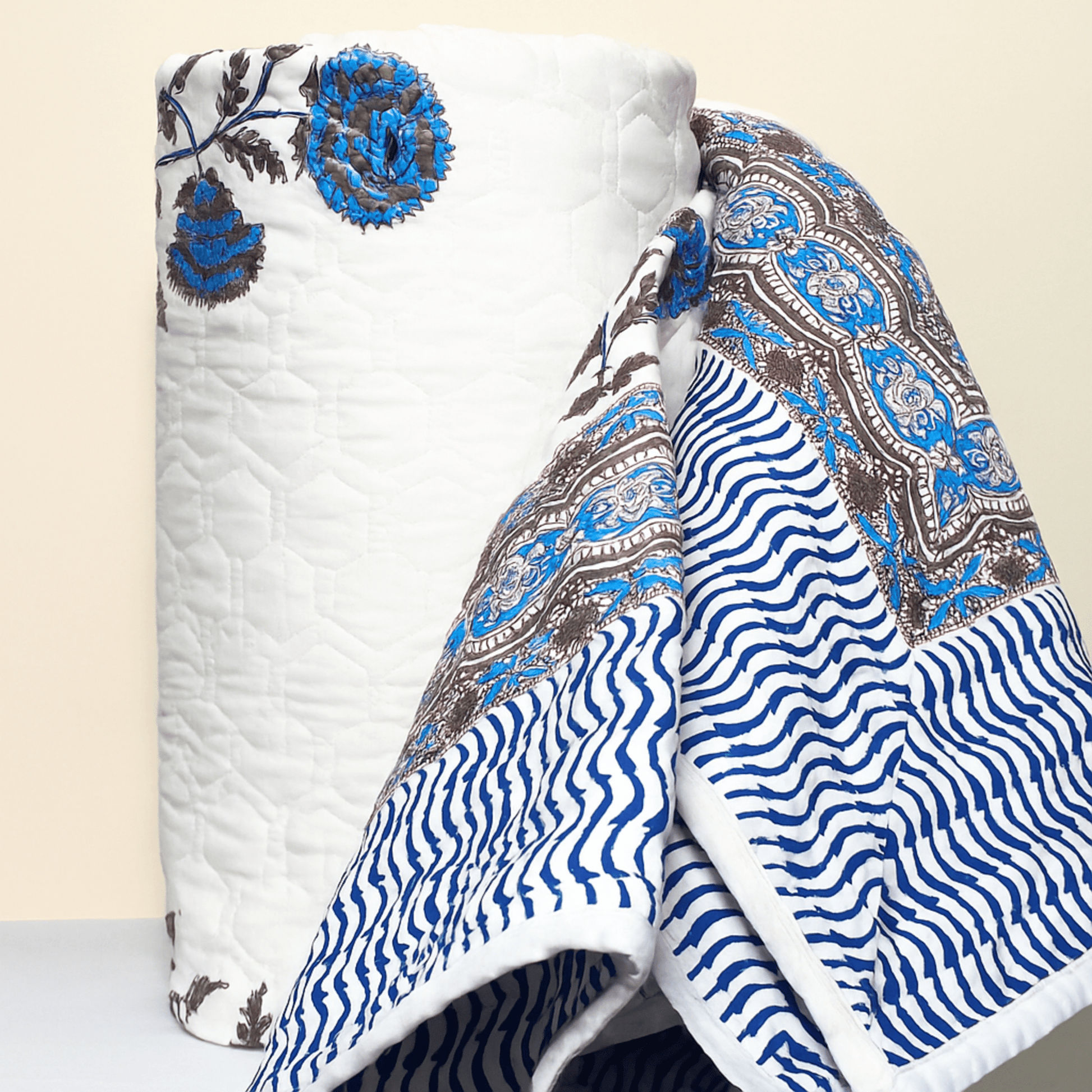 Rolled cotton quilt with handprinted blue flowers and intricate brown border, partially unrolled to reveal the reversible striped design.