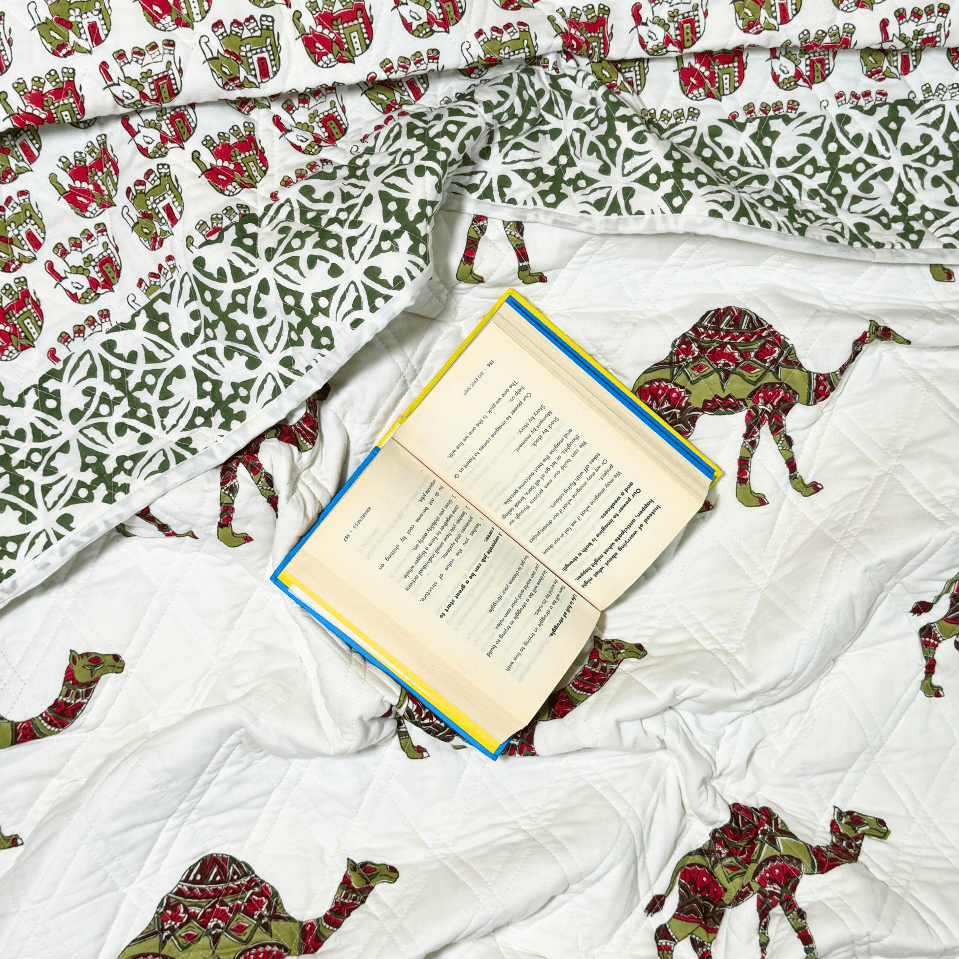 Flat lay of quilt with camels and palatial motifs, featuring a green printed border and an open book resting on the fabric.
