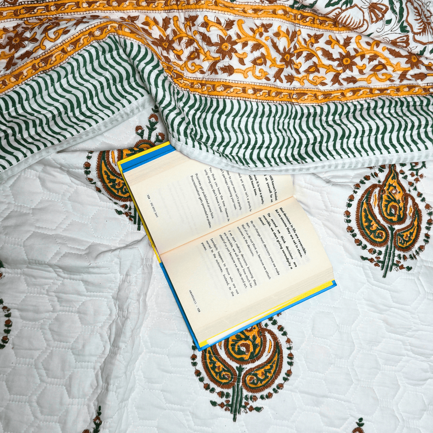 Flat lay of paisley handprinted quilt with wavy green borders and a book placed casually for a cozy reading setting.
