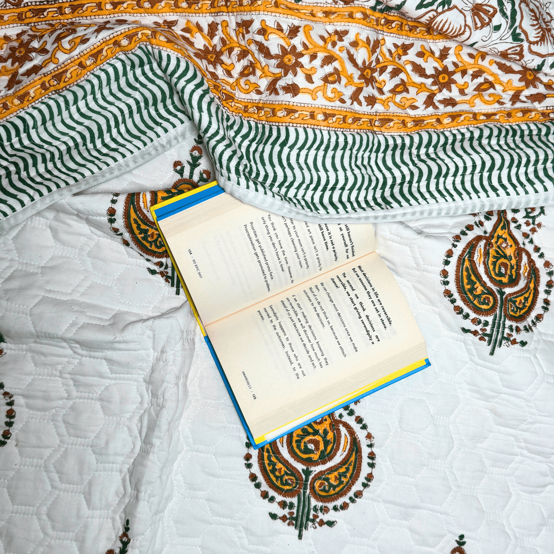Flat lay of paisley handprinted quilt with wavy green borders and a book placed casually for a cozy reading setting.
