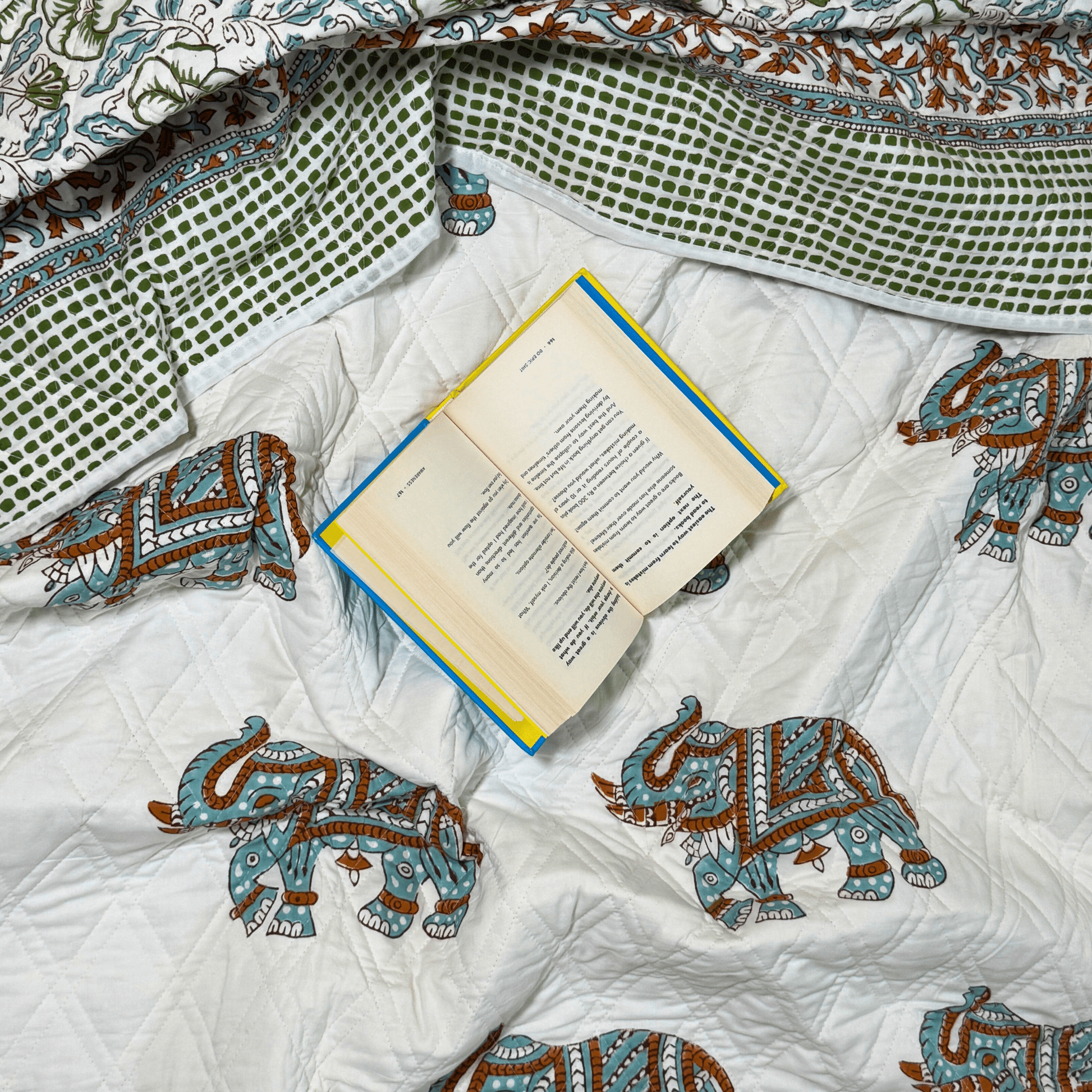 Flat lay of quilt featuring decorative elephants and leafy borders, with an open book placed casually for a cozy look.